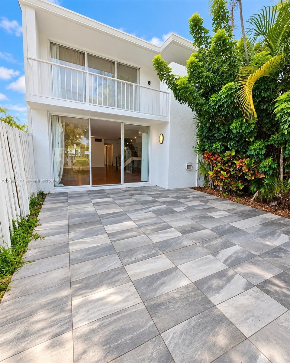 Exterior patio featuring large-format stone pavers, white stucco facade, second-story balcony with metal railings, sliding glass door entry, and lush perimeter landscaping