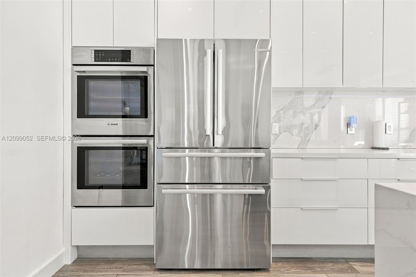 Kitchen featuring high-gloss white cabinetry, stainless steel double wall ovens, a stainless steel French-door refrigerator, stone-composite backsplash, and wood-finish flooring