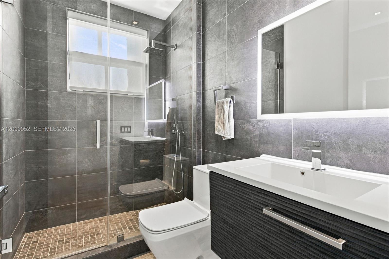 Bathroom featuring large-format dark grey wall tile, a glass-enclosed shower with a rainfall showerhead, a floating vanity with a textured cabinet finish, and a rectangular undermount sink with a backlit mirror