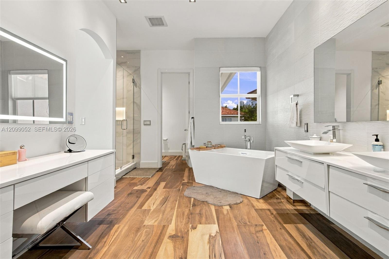 Wood-finish flooring, dual floating white vanities with vessel sinks, freestanding deep-soaking tub, and walk-in glass-enclosed shower