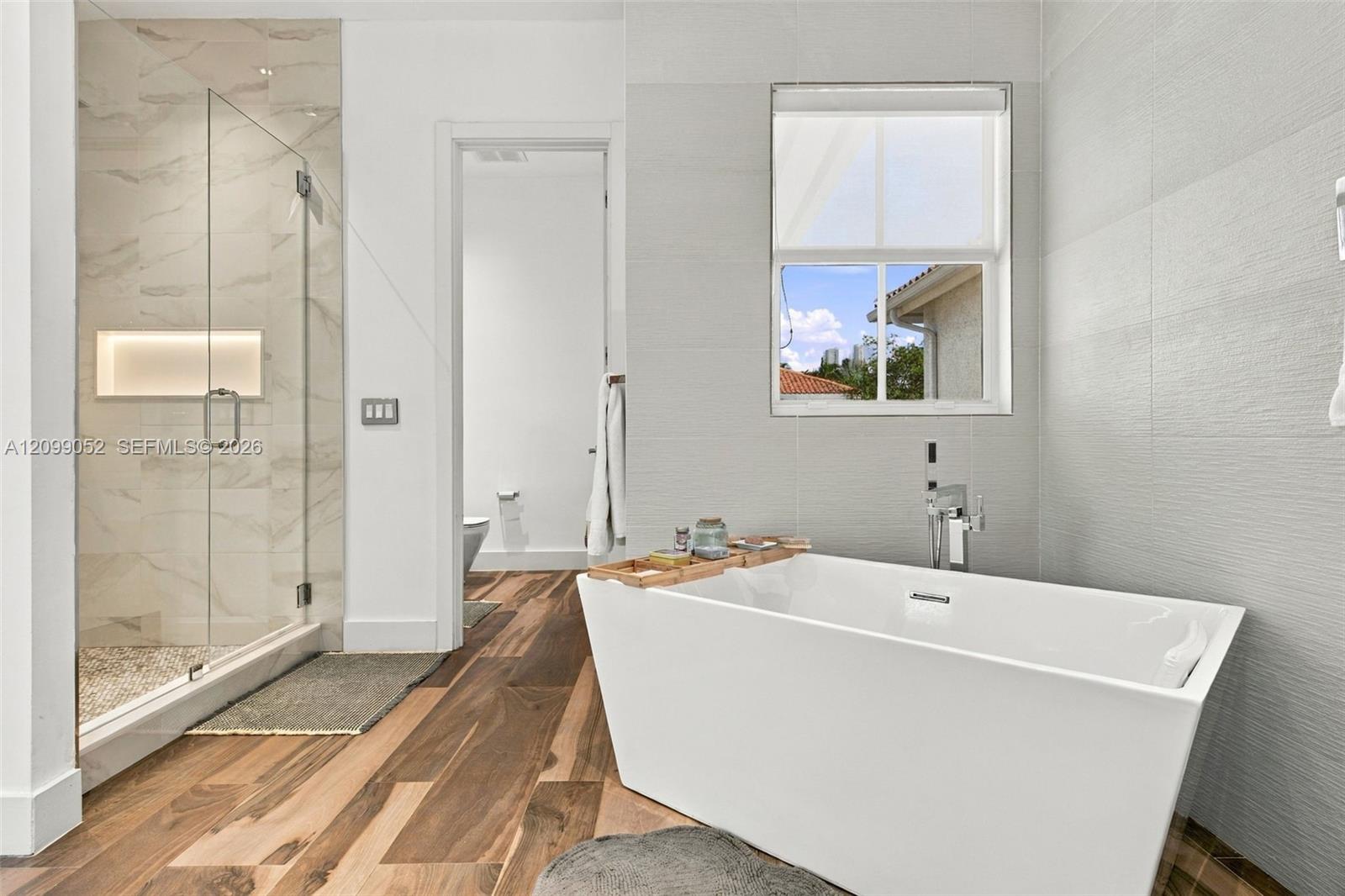 Freestanding soaking tub with chrome wall-mounted faucet, glass-enclosed shower with tiled walls and built-in recessed niche, and wood-finish flooring