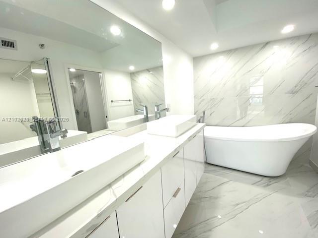 Bathroom featuring a dual-sink vanity with white rectangular vessel sinks, floor-to-ceiling veined stone-finish wall tile, a freestanding soaking tub, and matching veined stone-finish flooring
