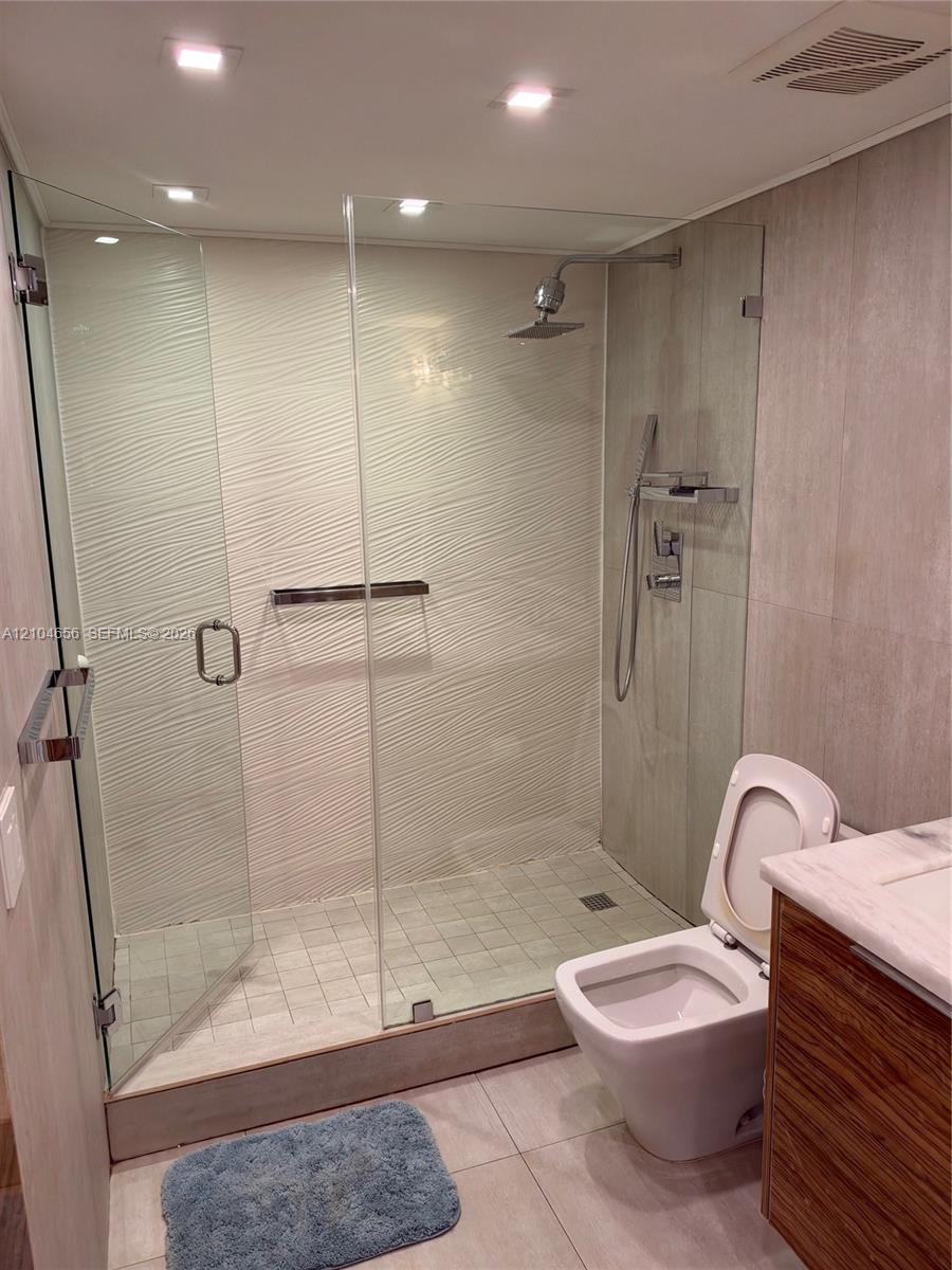 Walk-in shower featuring a glass enclosure, textured wall tile, ceiling-mounted rainfall showerhead, adjustable handheld shower wand, and tiled flooring