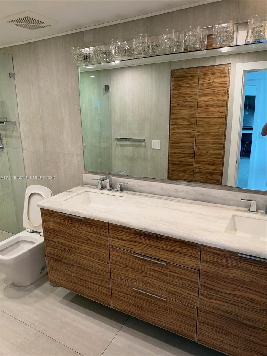 Double vanity with wood-finish cabinetry, integrated stone-surface countertop, dual rectangular undermount sinks, and a large-format mirror with multi-bulb overhead lighting