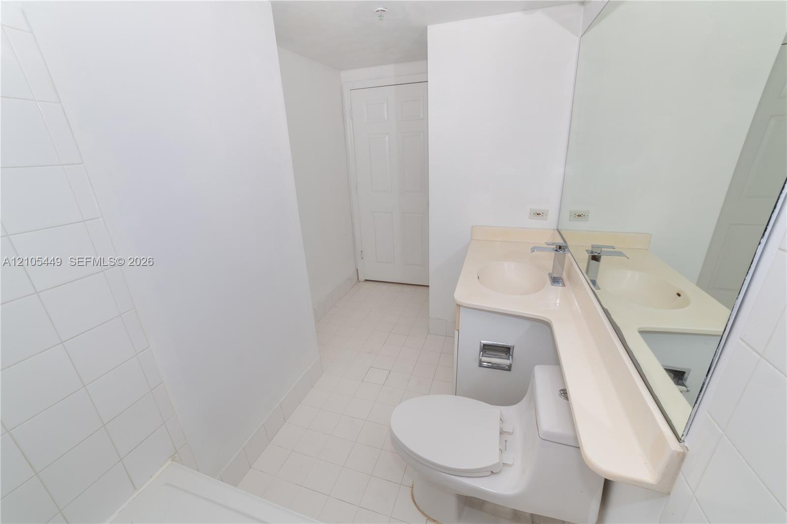 White tile flooring, white tile shower surround, integrated sink vanity with a light-colored countertop, and a large wall-mounted mirror