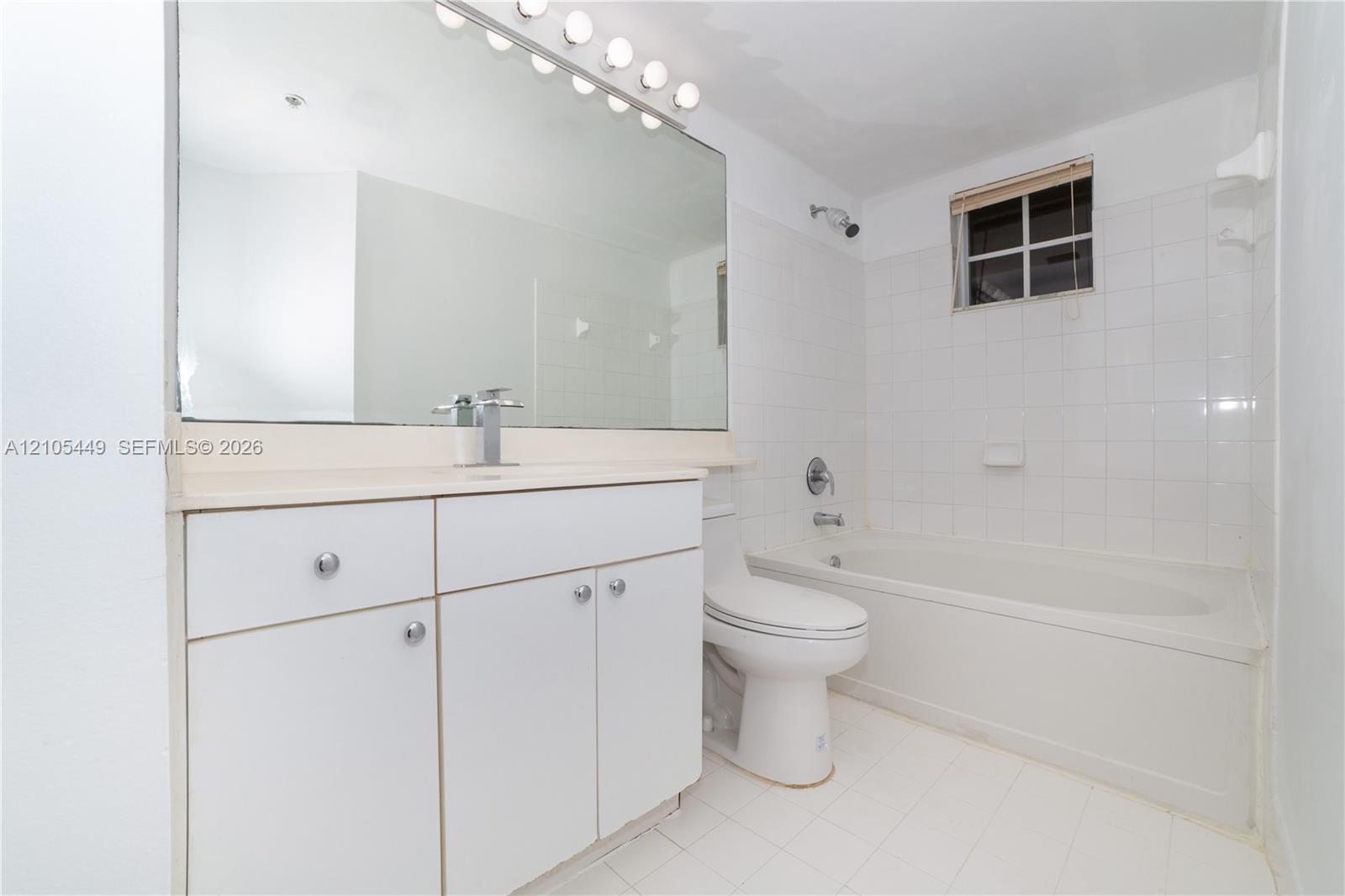 White tile flooring, white vanity with integrated sink, chrome faucet, wall-mounted mirror with overhead lighting, and built-in bathtub with white tile surround