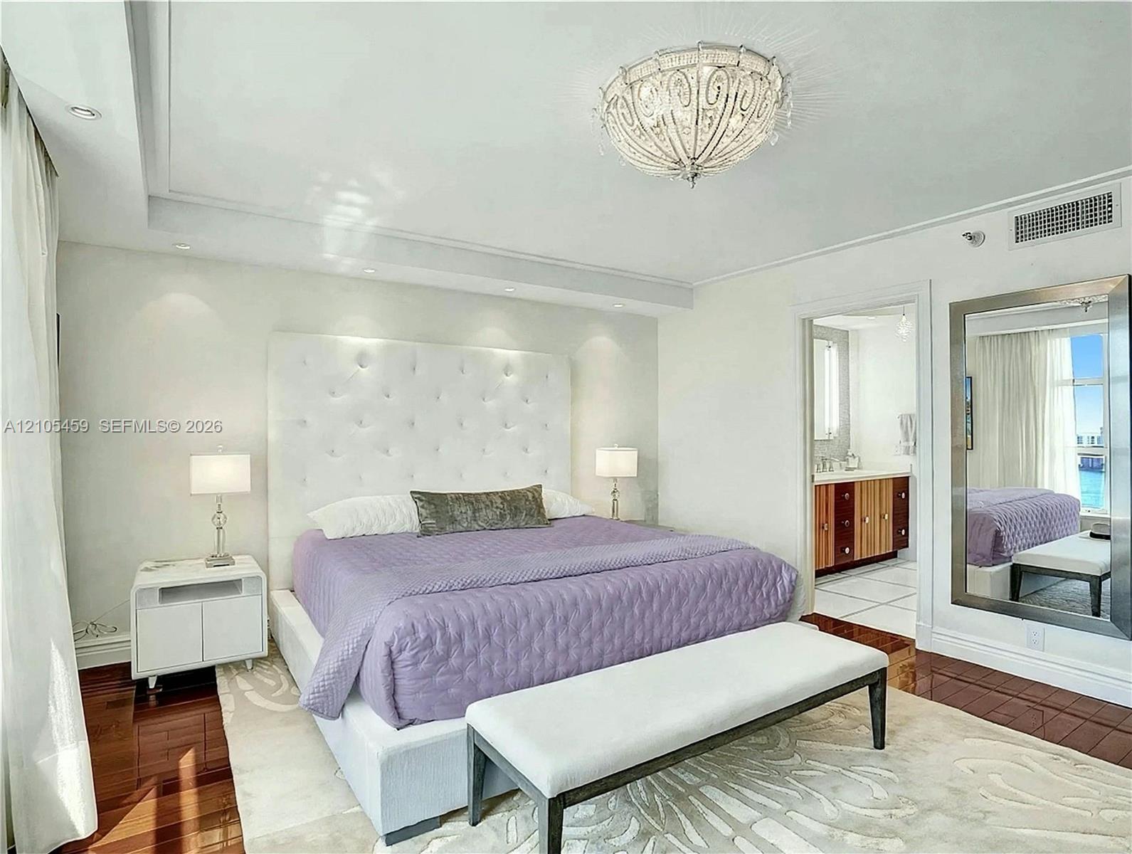 Tray ceiling with recessed lighting, ornate ceiling fixture, wood-finish flooring, tufted headboard, and an en-suite bathroom with a wood-finish vanity