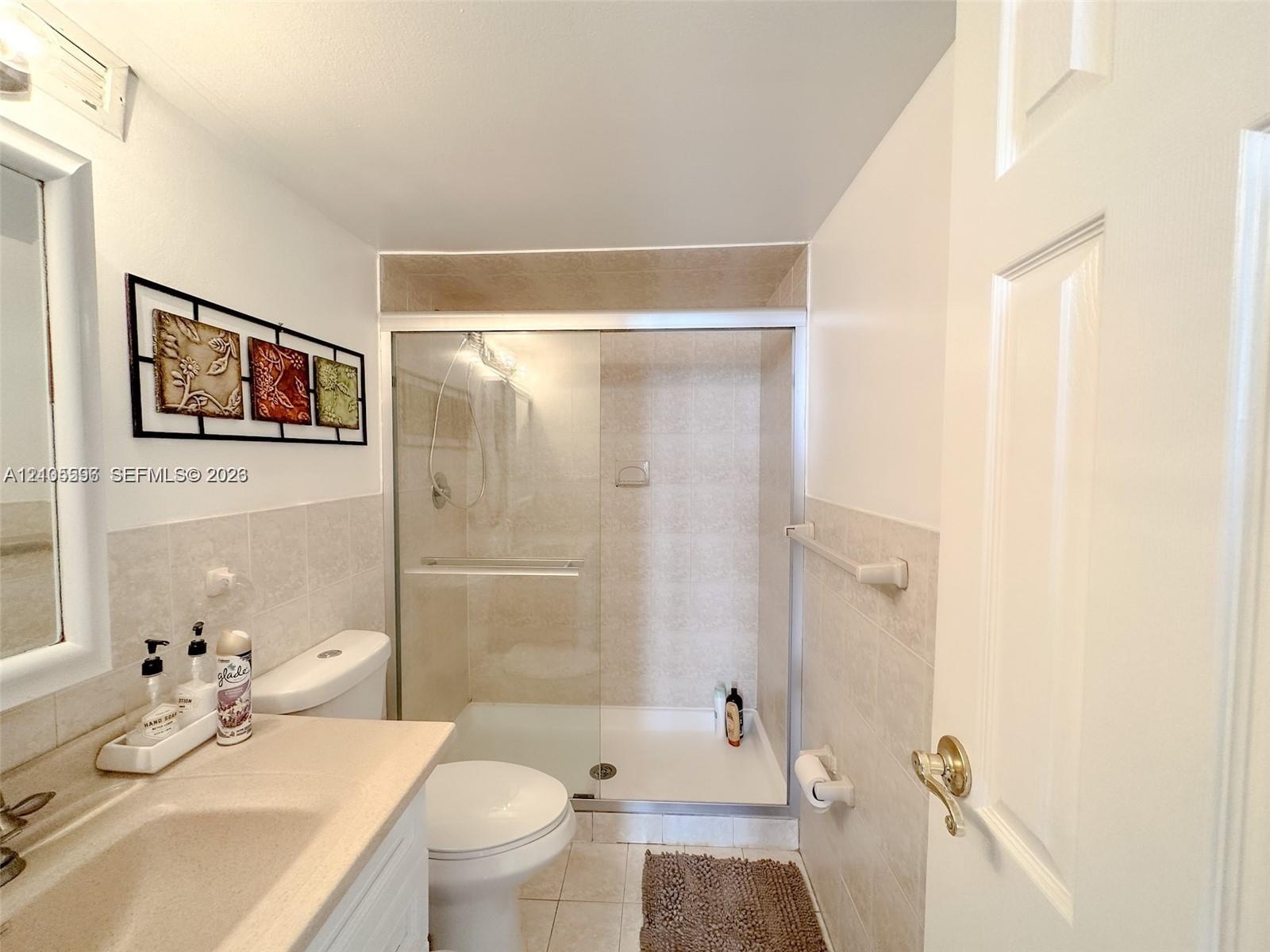 Bathroom featuring a walk-in shower with glass sliding doors, ceramic tile shower surround, neutral solid-surface vanity countertop, and ceramic floor tiling