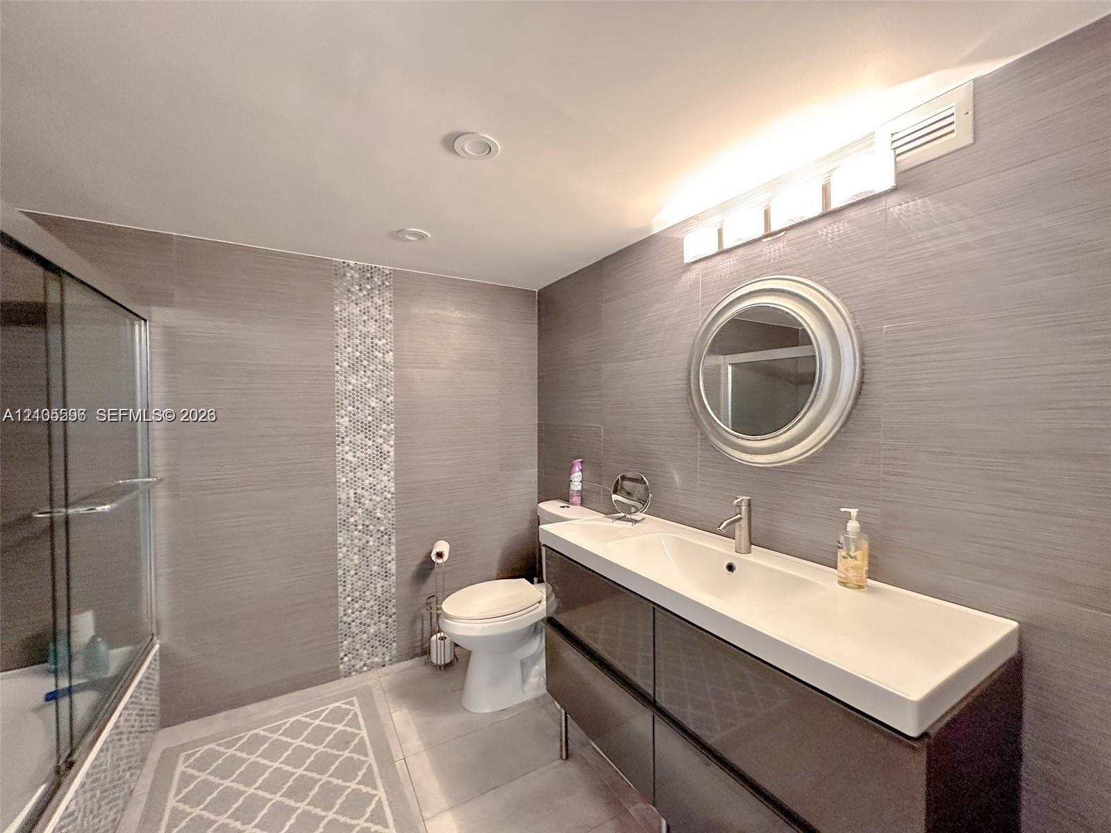 Bathroom featuring gray wall tile with vertical mosaic accent inlay, a wall-mounted vanity with integrated sink, a framed circular mirror, and a glass-enclosed shower