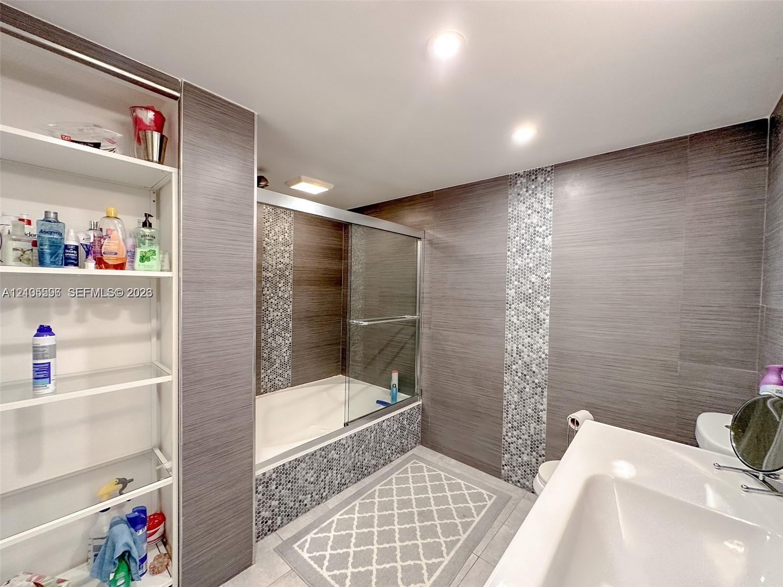 Bathroom featuring large-format horizontal gray wall tiling, vertical glass mosaic tile accents, a built-in recessed shelving unit, a glass-enclosed bathtub, and recessed ceiling lighting