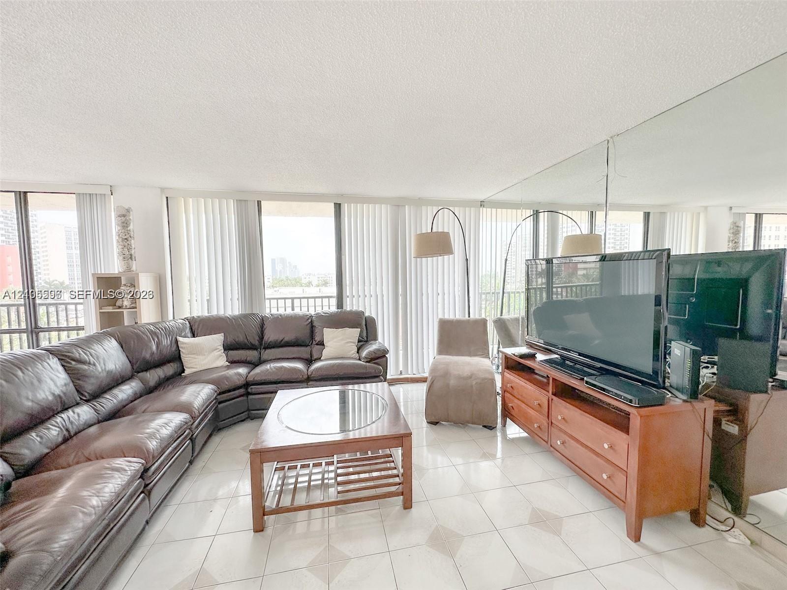 Expansive living area featuring light-toned tile flooring, floor-to-ceiling vertical blinds, and a large wall-mounted mirror