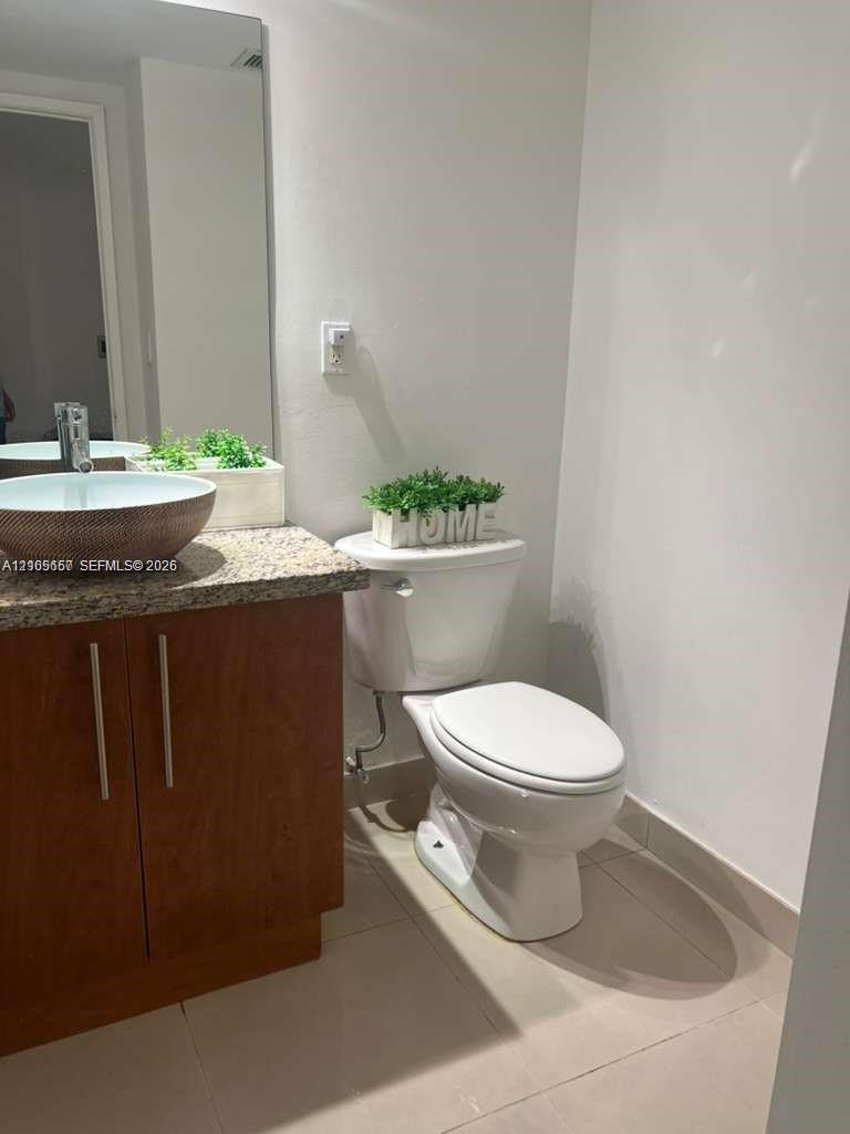 Wood-finish cabinetry, stone-texture countertop, vessel sink with metal faucet, large wall-mounted mirror, and tile flooring