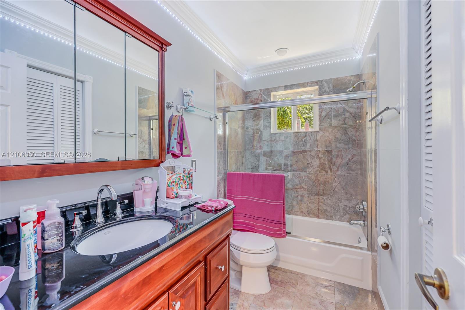 Bathroom featuring a wood-finish vanity with a dark polished countertop, undermount sink, multi-panel medicine cabinet, and a bathtub enclosure with stone-patterned tile surround