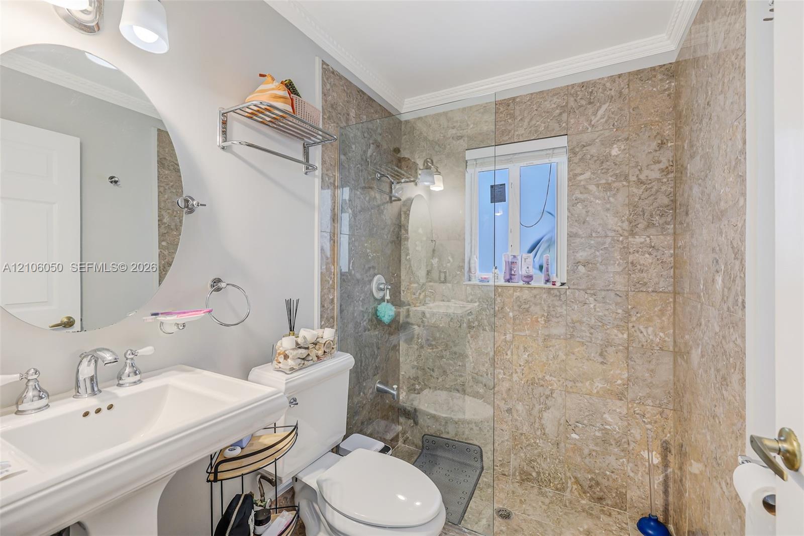Bathroom featuring a wall-mounted oval mirror, porcelain pedestal sink, chrome hardware, and a glass-enclosed walk-in shower finished with full-height stone-patterned wall tile