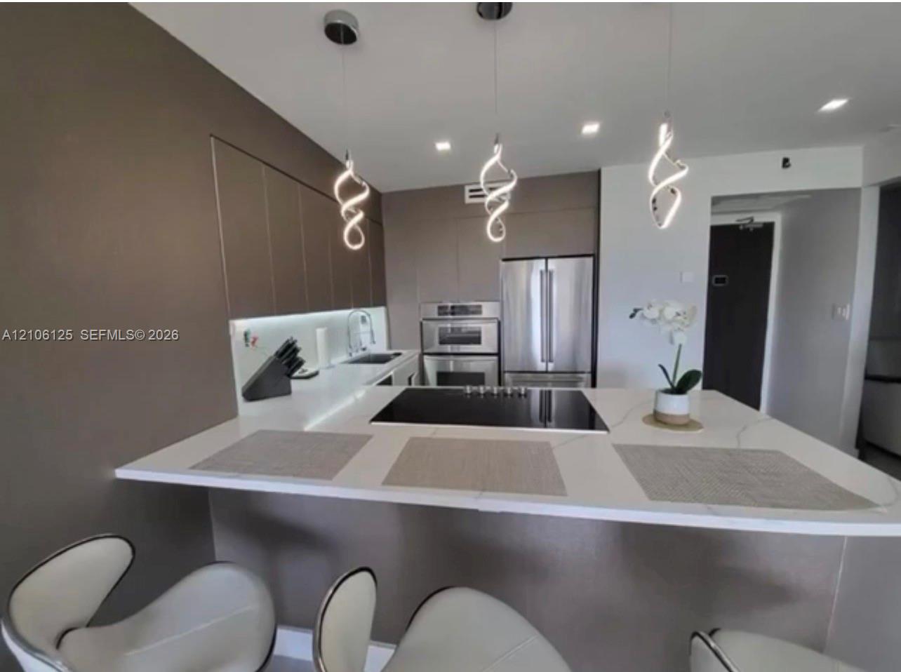 Contemporary kitchen featuring a spacious quartz peninsula, stainless steel double wall ovens, a flat-surface cooktop, matte-finish cabinetry, and sculptural pendant lighting