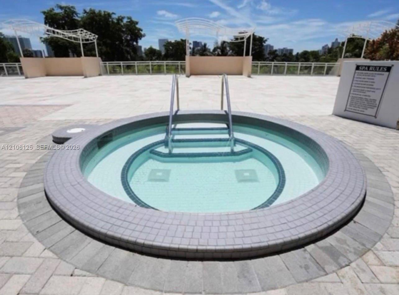 Circular spa featuring tiled surround, metal handrail, paver hardscaping, and overhead pergola structures