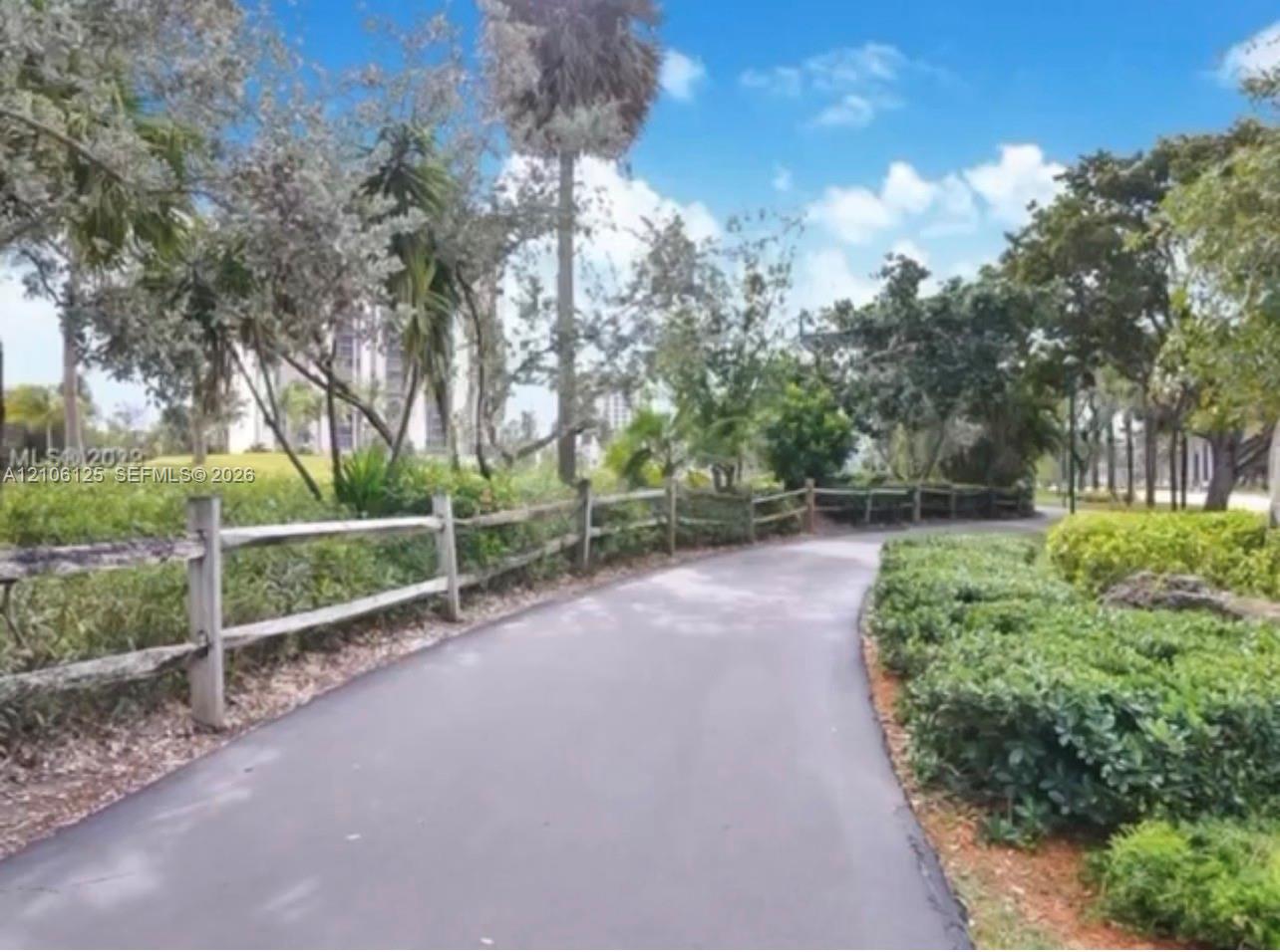 Paved pathway bordered by split-rail fencing, mature trees, and established shrubbery