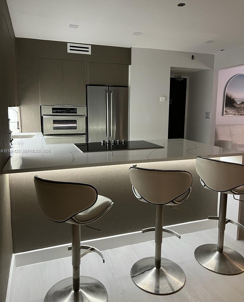 Kitchen featuring flat-panel cabinetry, stainless steel appliances including a wall-mounted oven, and a solid-surface waterfall island with an integrated cooktop and under-counter lighting