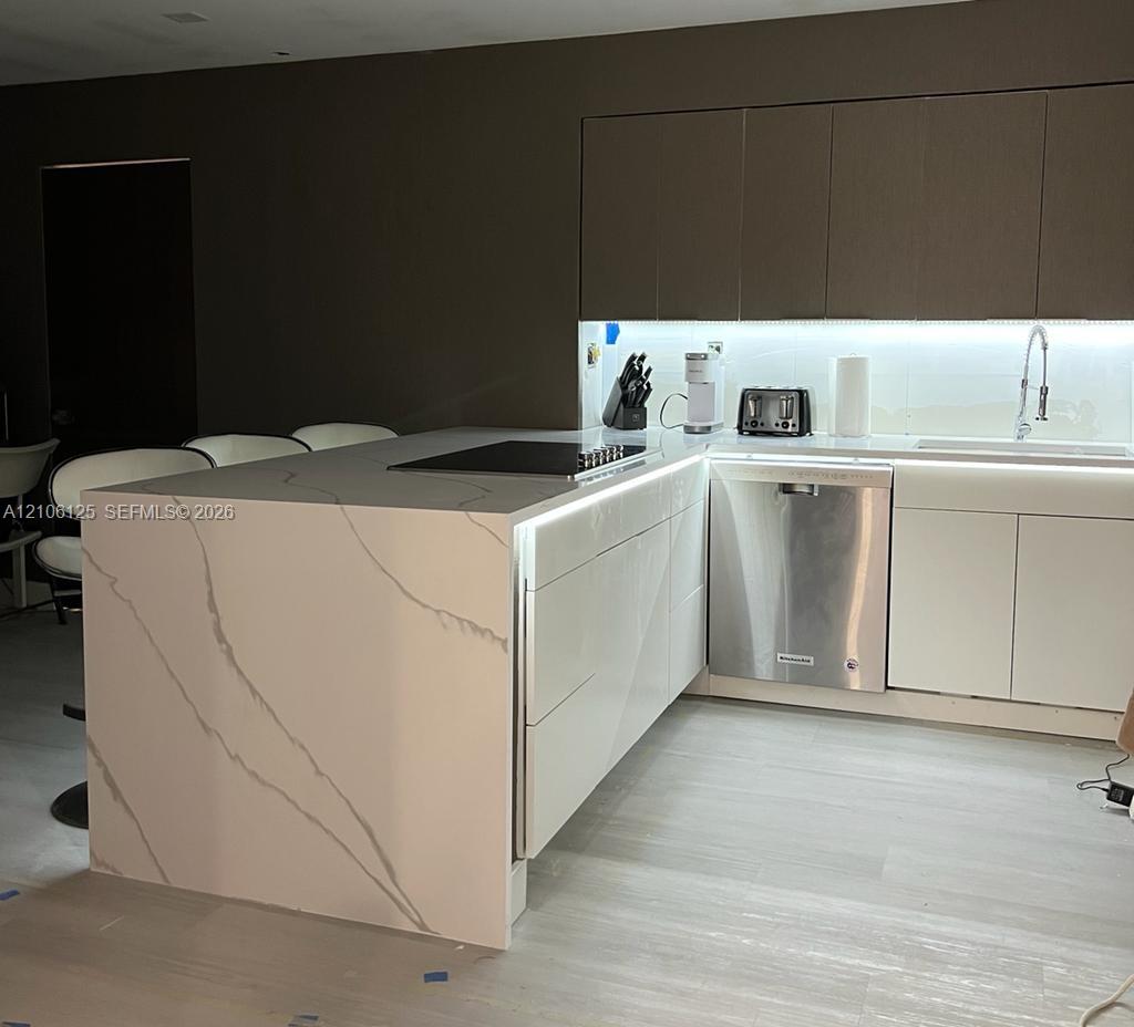 Contemporary kitchen featuring a stone waterfall island with an integrated cooktop, flat-panel cabinetry, stainless steel dishwasher, and under-cabinet accent lighting