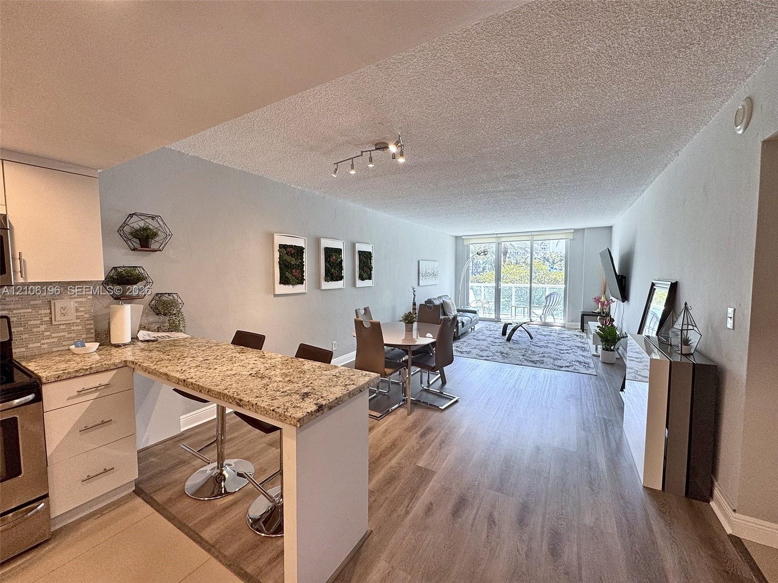 Open-concept living space featuring wood-finish flooring, stone-surfaced breakfast bar, stainless steel appliances, track lighting, and floor-to-ceiling glass sliding doors