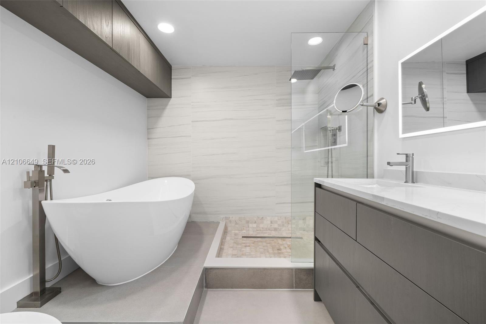Freestanding soaking tub with floor-mounted faucet, walk-in glass-enclosed shower featuring mosaic stone flooring, dark wood-finish cabinetry, illuminated wall-mounted mirror, and recessed lighting