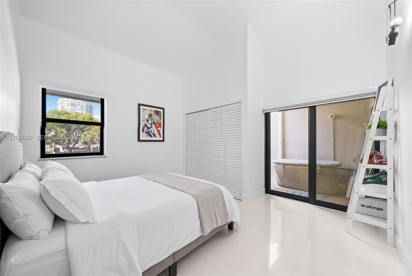 High-ceiling room featuring white tile flooring, louvered-panel closet doors, and a large sliding glass door with access to an exterior area