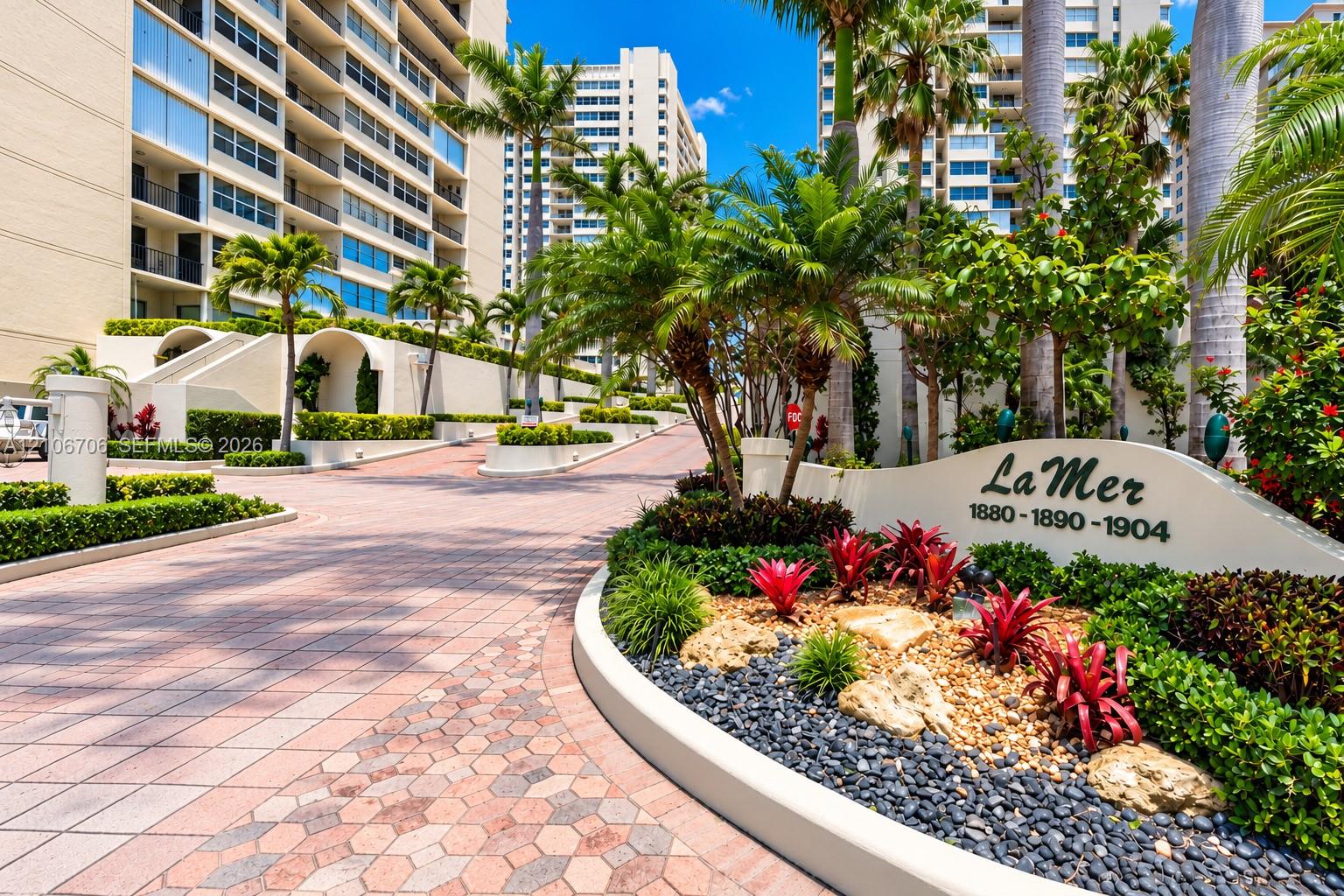 Paver driveway with geometric detailing, manicured tropical landscaping, mature palm trees, multi-story masonry building exterior, and decorative stone accent signage