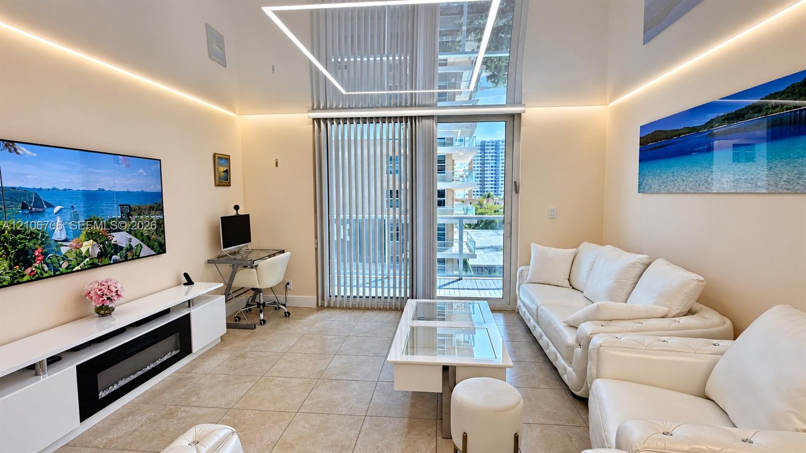 Living area featuring high-gloss ceiling panels with perimeter LED accent lighting, integrated electric fireplace, and full-height sliding glass doors with vertical blinds