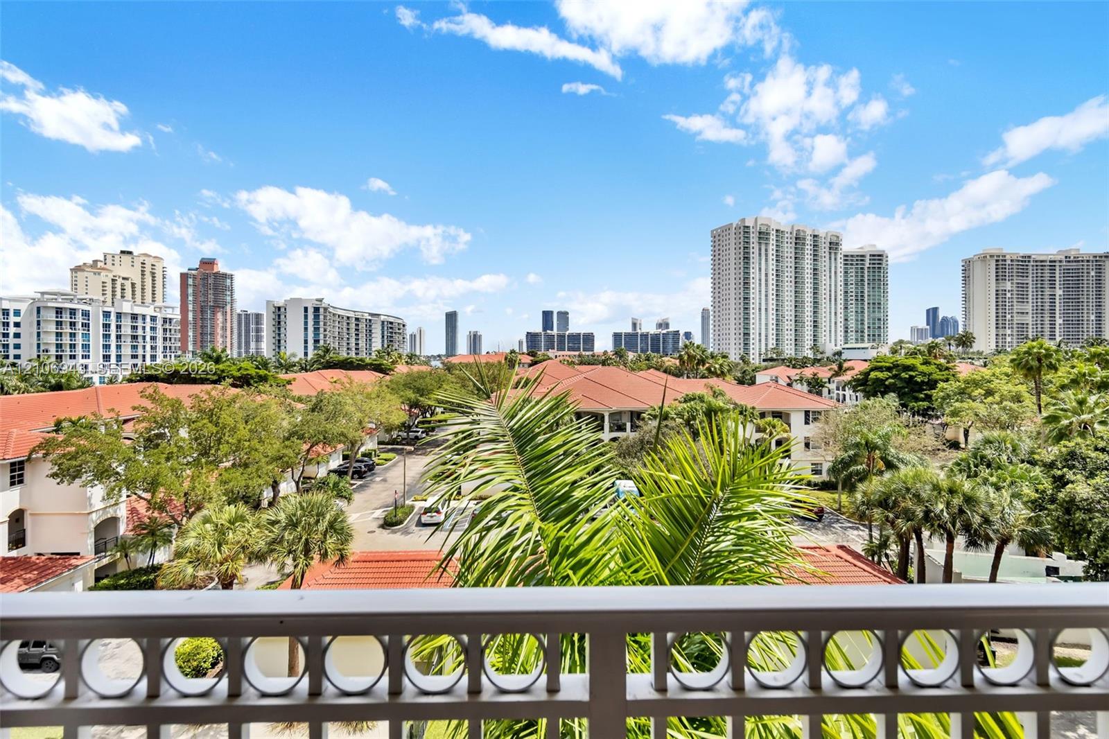 Elevated balcony featuring a decorative metal railing, overlooking a cityscape with terracotta rooflines and mature palm trees