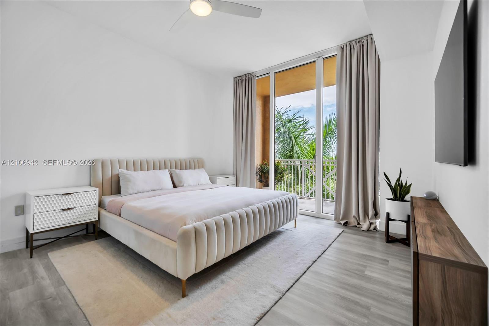 Floor-to-ceiling glass sliding doors opening to a balcony, light-toned wood-finish flooring, and a ceiling-mounted light fixture with integrated fan