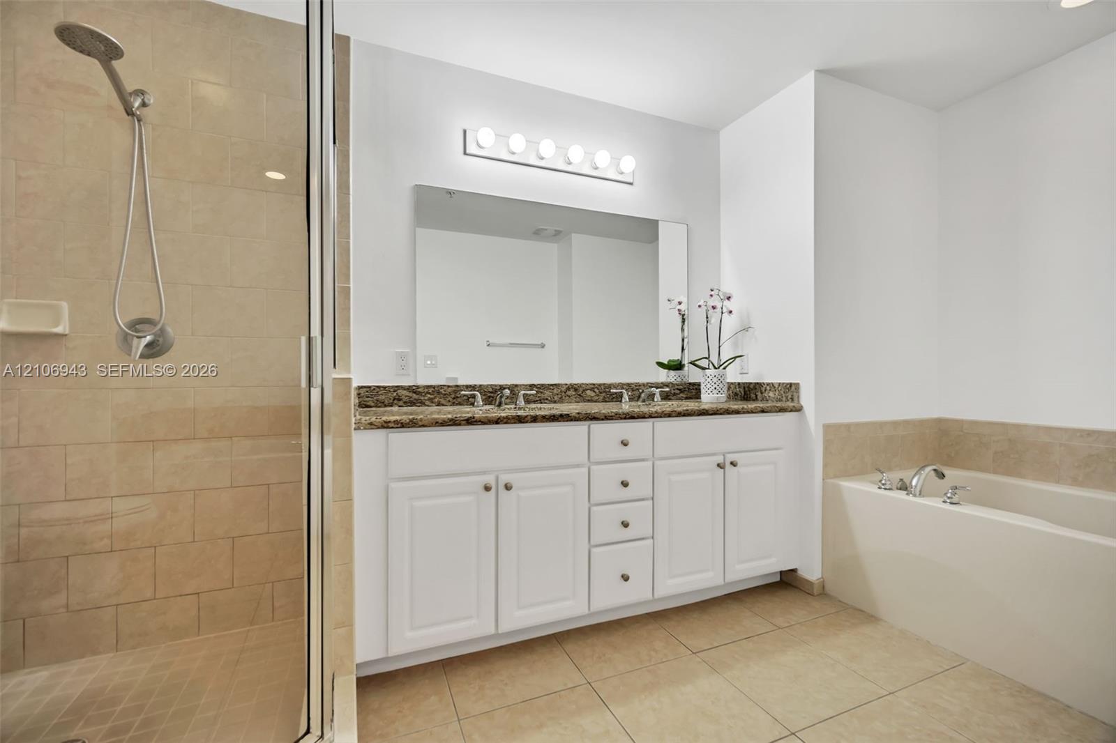 Double vanity with stone countertop, white shaker-style cabinetry, walk-in tile shower with handheld showerhead, built-in soaking tub, and wall-mounted multi-light fixture