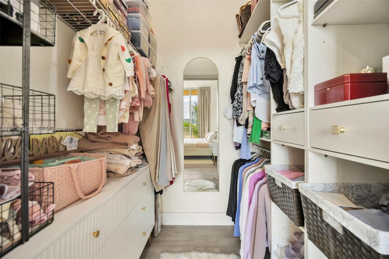 Walk-in closet featuring white built-in cabinetry with integrated drawers, brass hardware, wire shelving, and a full-length arched mirror
