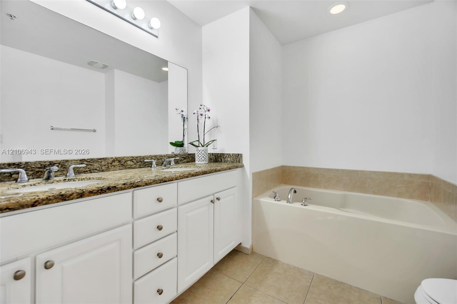 Dual-sink vanity featuring stone-finish countertop, white cabinetry with integrated storage, full-width wall mirror, and a built-in soaking tub with surround tile