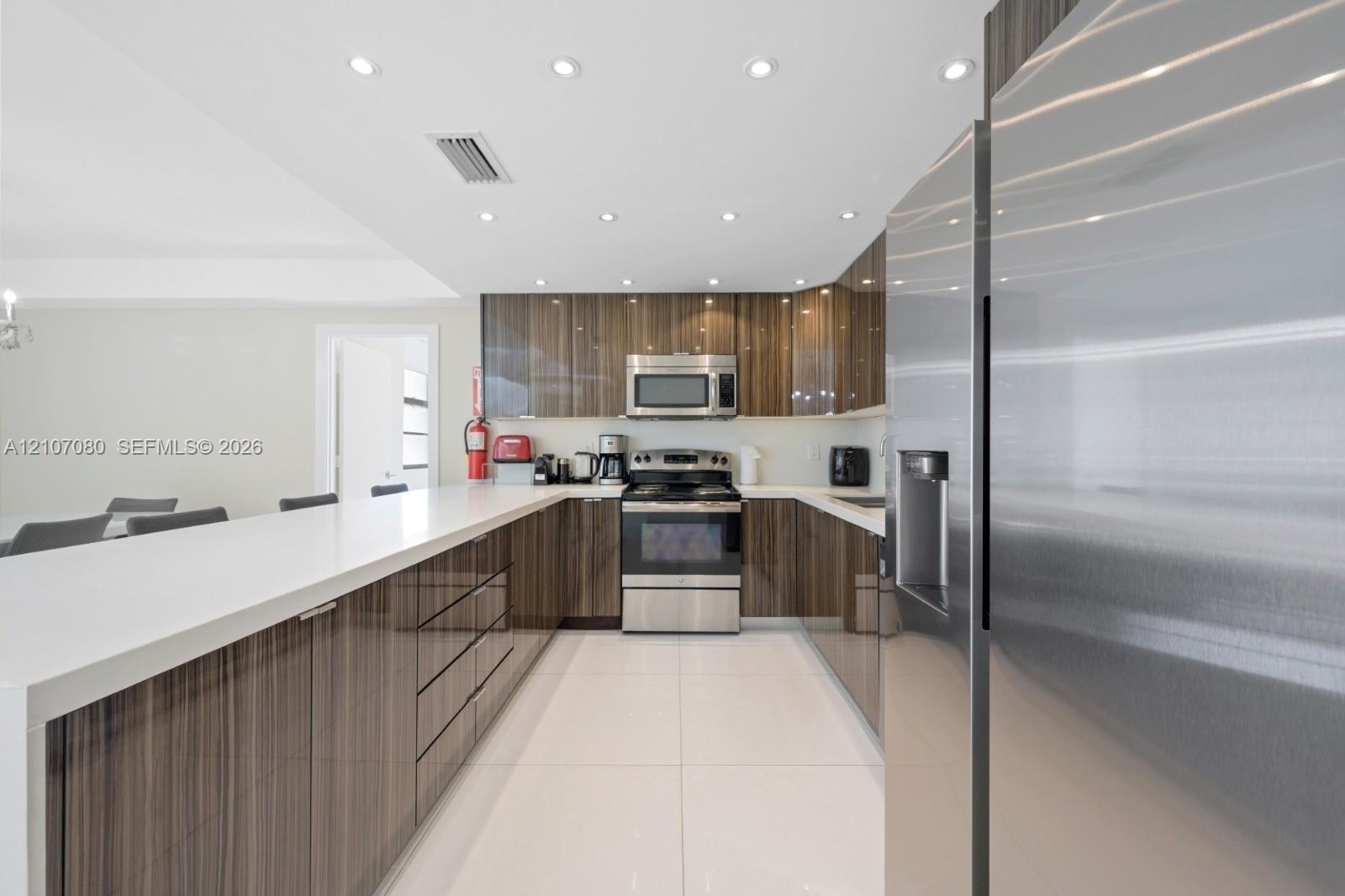 Kitchen featuring high-gloss wood-finish cabinetry, white solid-surface countertops, large-format tile flooring, stainless steel appliances, and recessed ceiling lighting