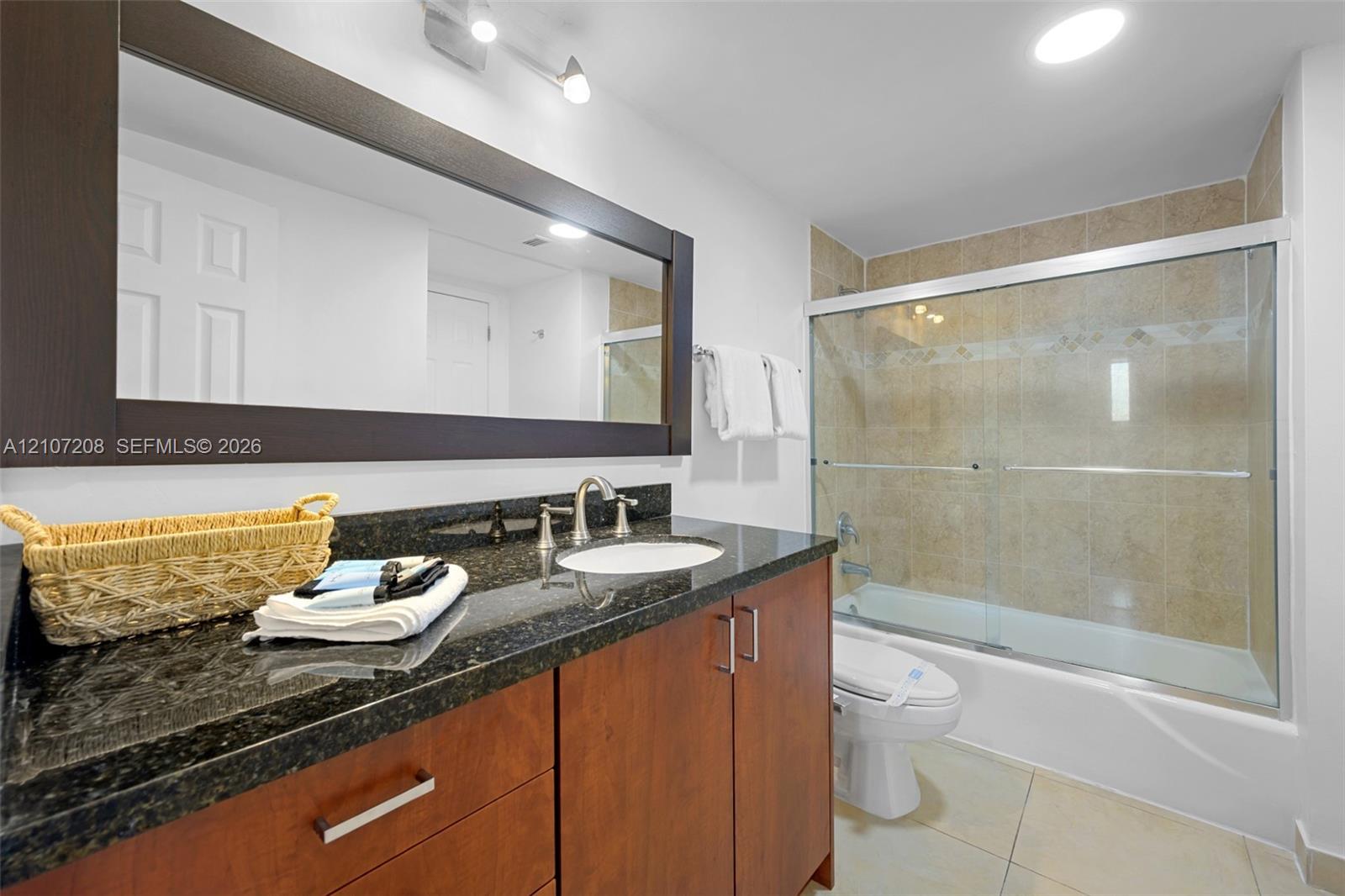 Bathroom featuring a wood-finish vanity with a dark stone countertop, a large framed wall mirror, and a tub-shower combination with tiled surround and glass sliding doors