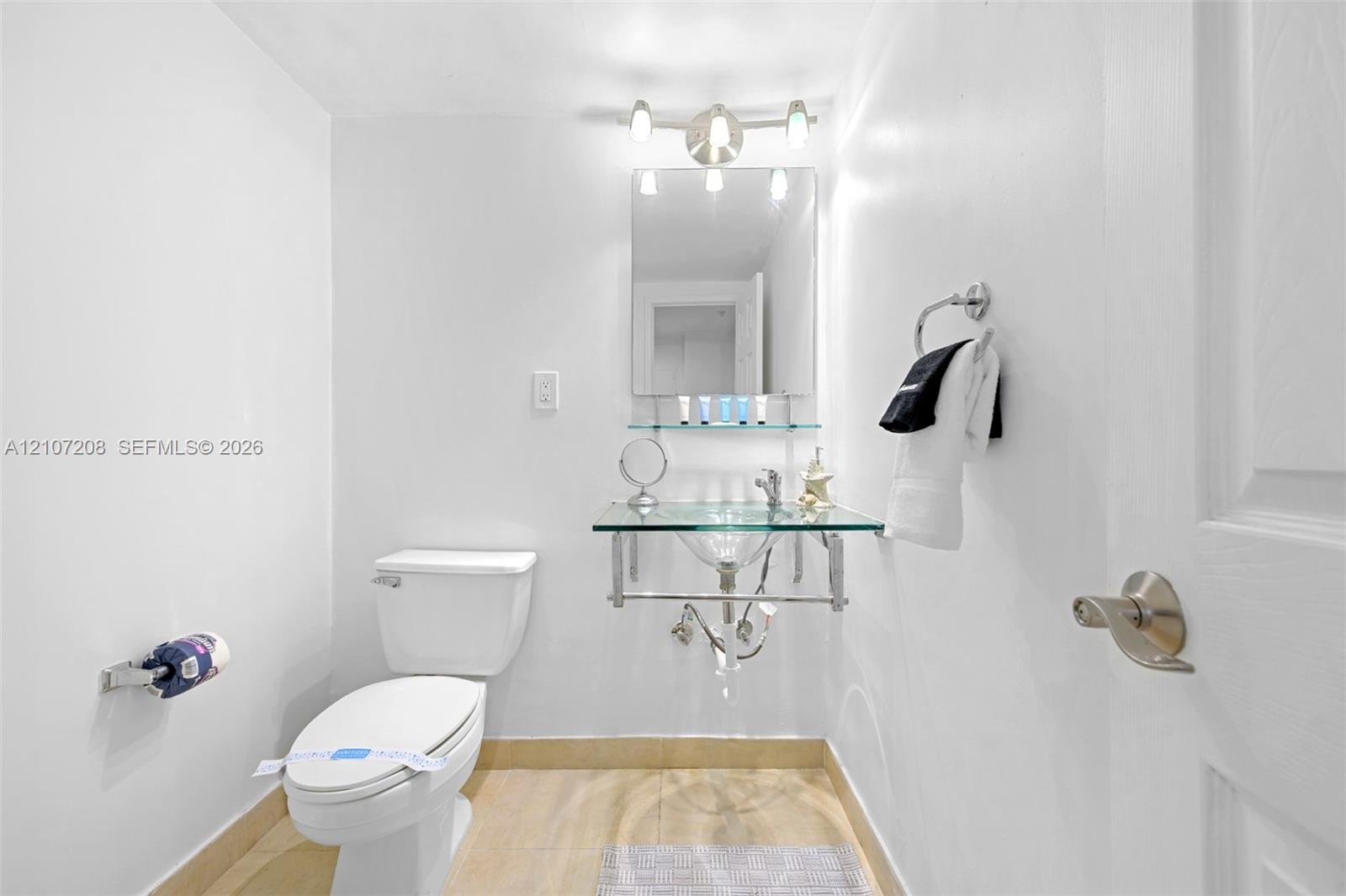Glass-top wall-mounted sink with chrome hardware, rectangular mirror, glass shelf, three-light vanity fixture, and porcelain-finish flooring