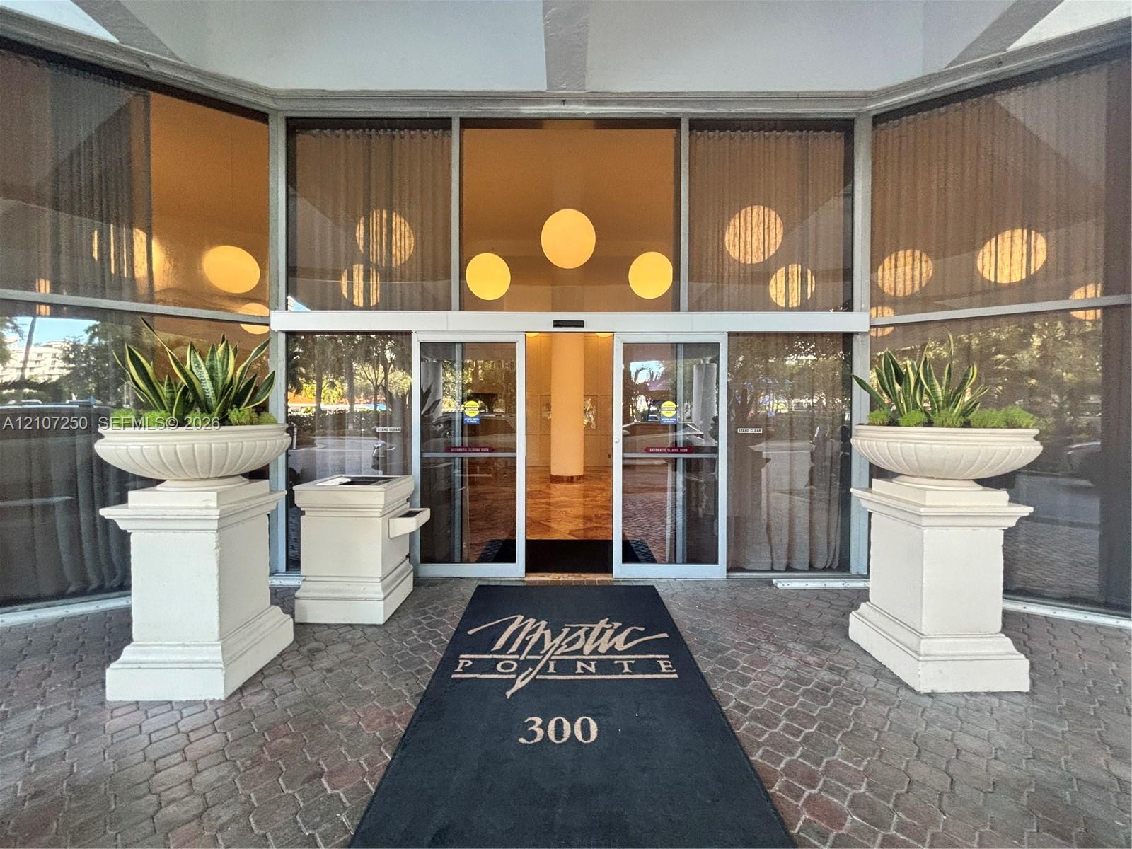 Grand entrance featuring expansive floor-to-ceiling glass paneling, automatic sliding glass doors, and dual oversized stone-finish pedestal planters