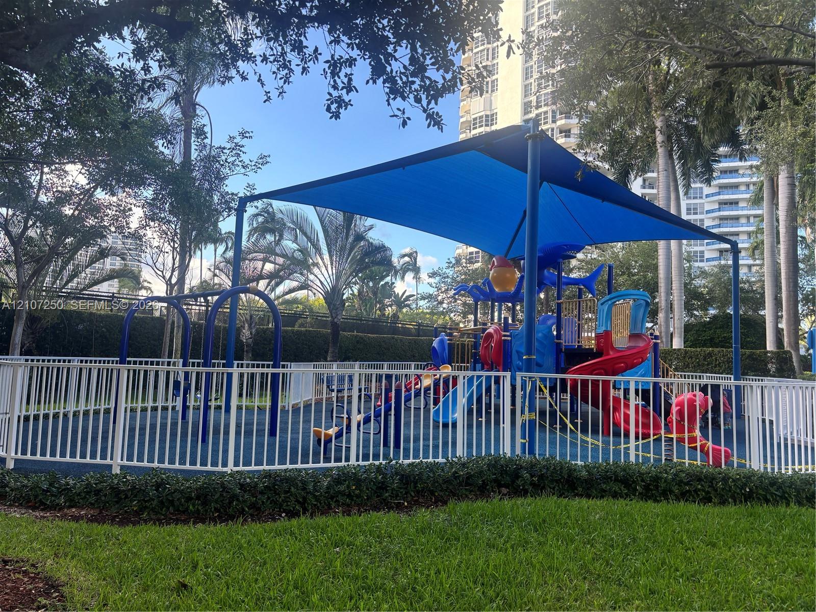Recreational area featuring a modular playground structure, overhead shade canopy, protective rubberized surface, and white perimeter fencing