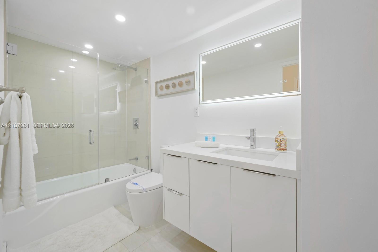 Bathroom featuring a white floating vanity with a solid-surface countertop, an integrated rectangular sink, a glass-enclosed shower-tub combination, and an LED-backlit mirror