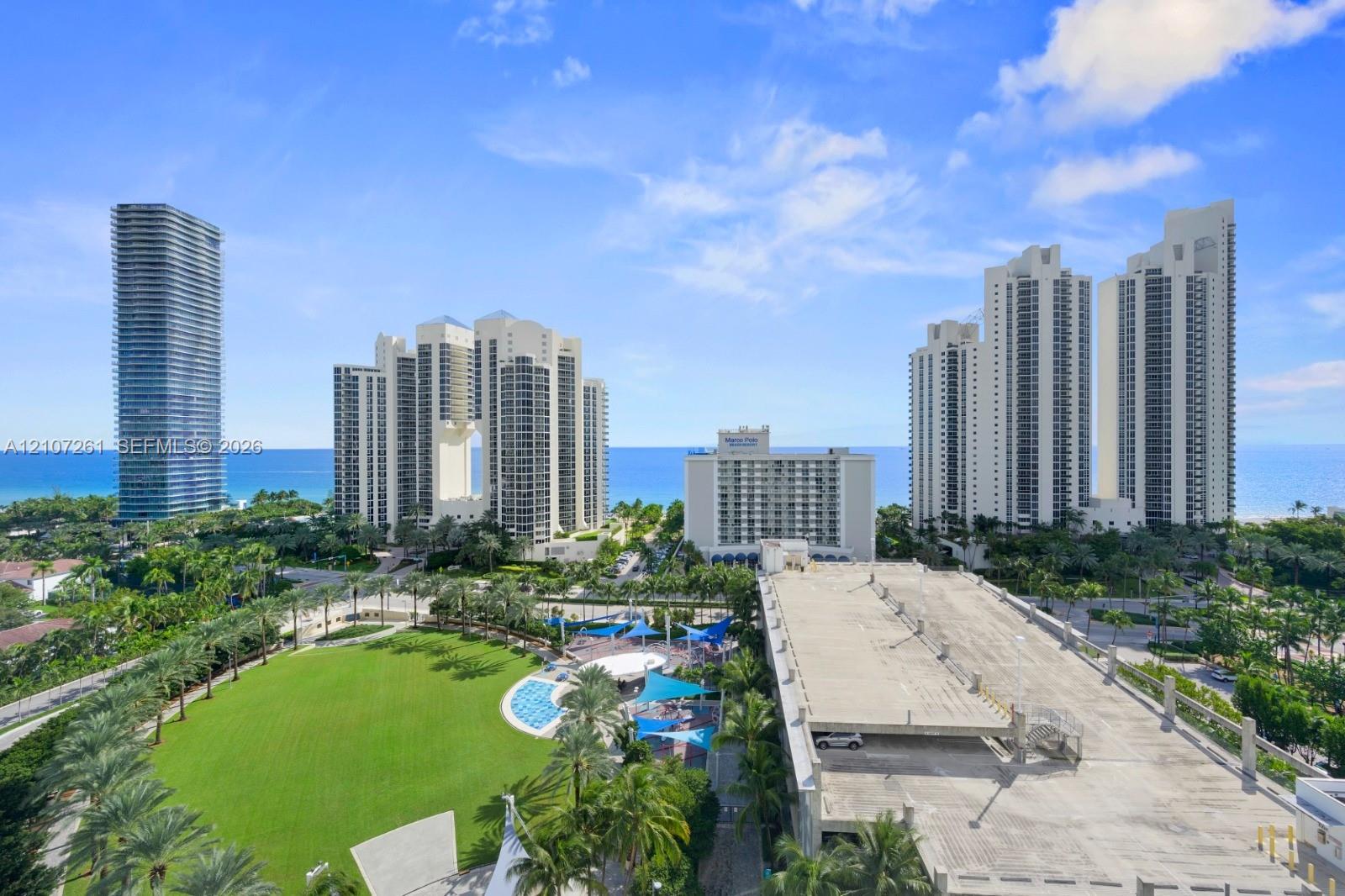 Aerial perspective featuring expansive green space, mature palm trees, multi-story high-rise architecture, and coastal proximity