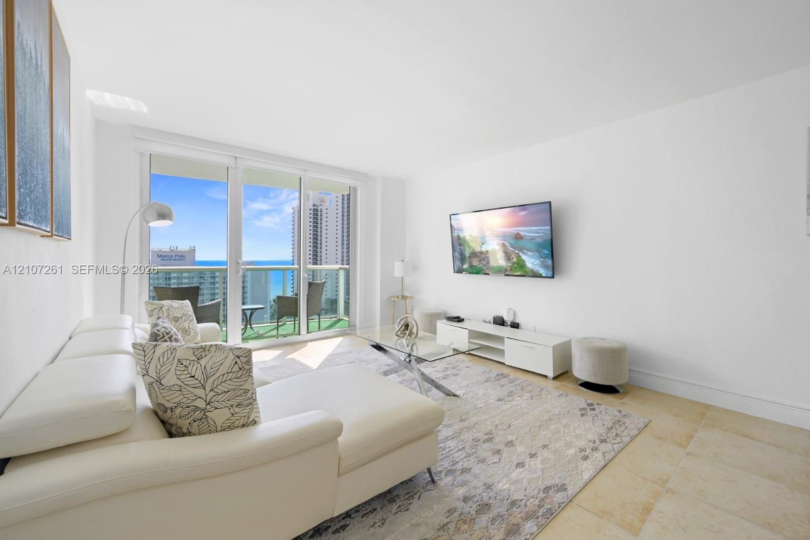 Living area featuring expansive floor-to-ceiling glass sliding doors, private balcony, and light-toned stone tile flooring