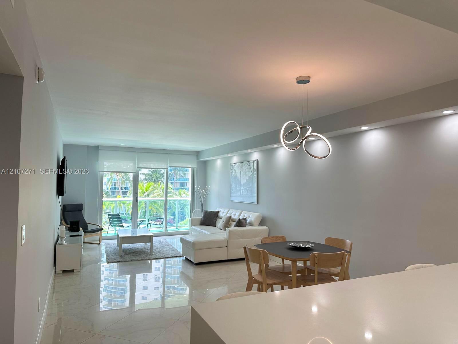 Open-concept area featuring polished tile flooring, recessed ceiling lighting, a modern sculptural chandelier, and a sliding glass door leading to a balcony with a glass railing
