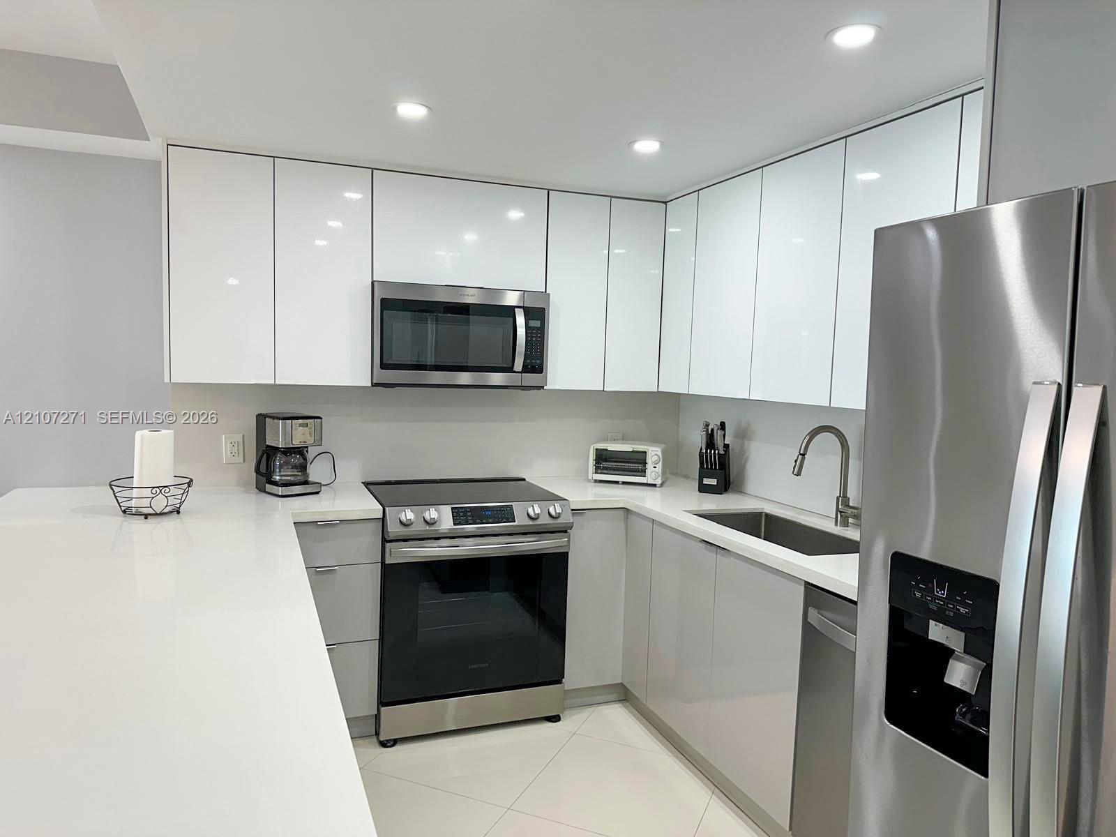 Modern kitchen featuring high-gloss white cabinetry, stainless steel appliances, a stone countertop, recessed ceiling lighting, and large-format tile flooring