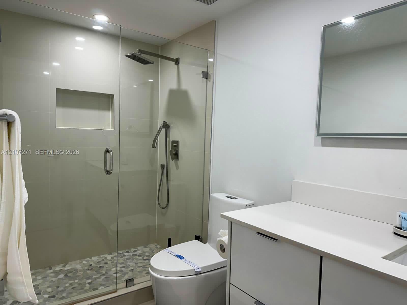 Bathroom featuring a frameless glass walk-in shower with a recessed wall niche, pebble-stone flooring, rainfall showerhead, and a vanity with a solid-surface countertop