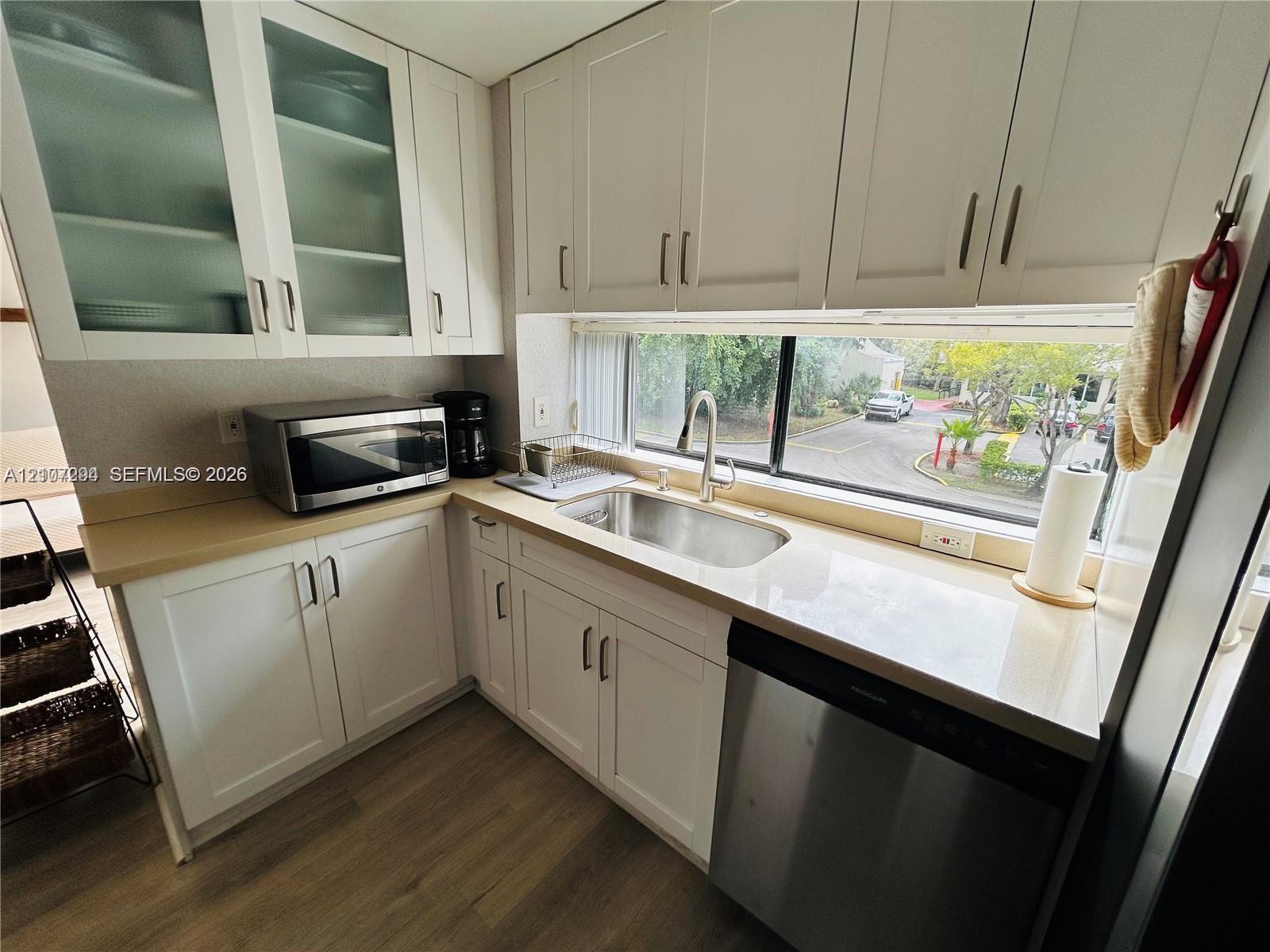 Kitchen featuring shaker-style cabinetry with glass-front accents, solid-surface countertops, undermount stainless steel sink, stainless steel dishwasher, and wood-finish flooring