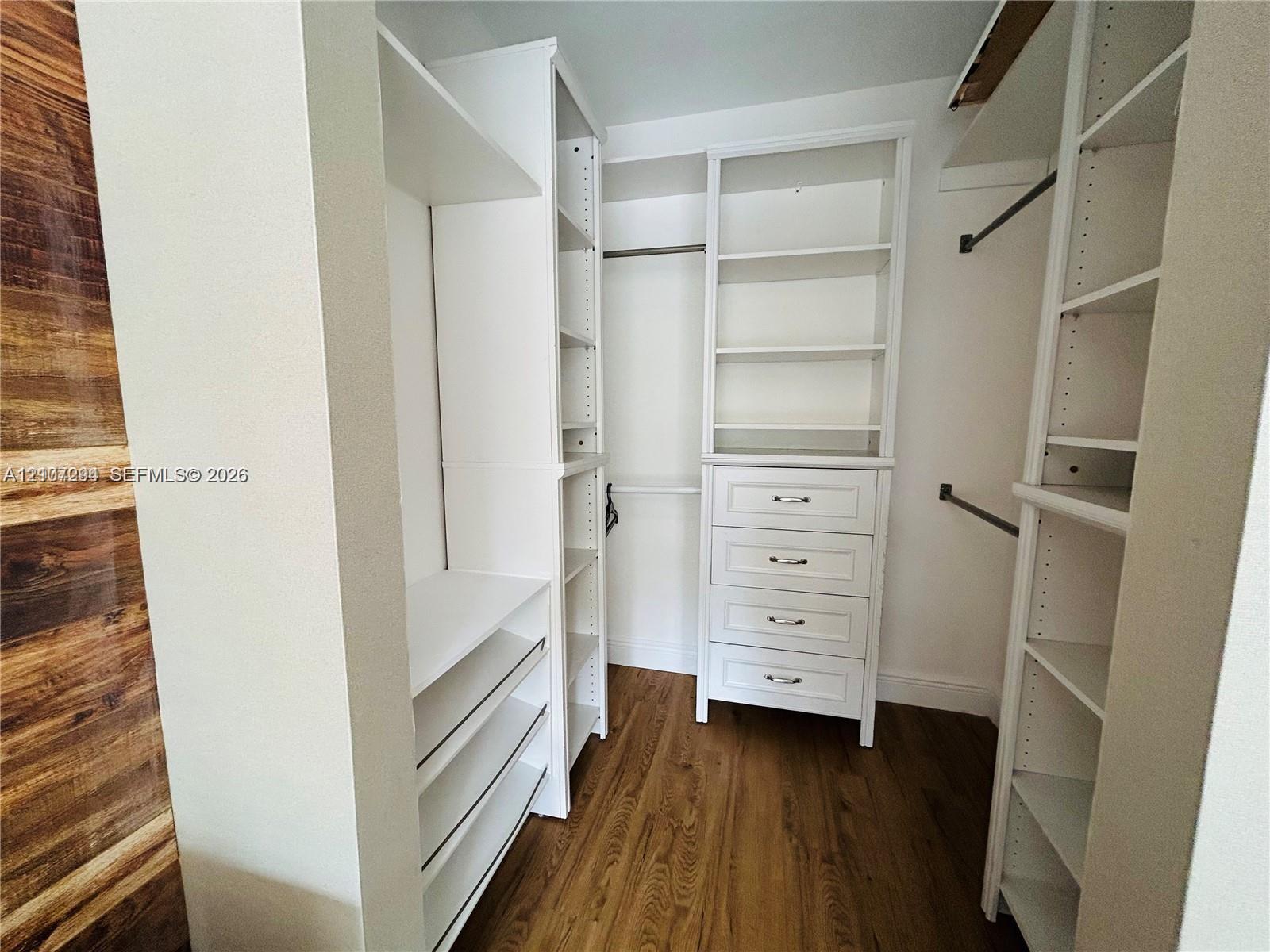 Walk-in closet featuring custom white built-in shelving, a four-drawer dresser unit, integrated hanging rods, and wood-finish flooring