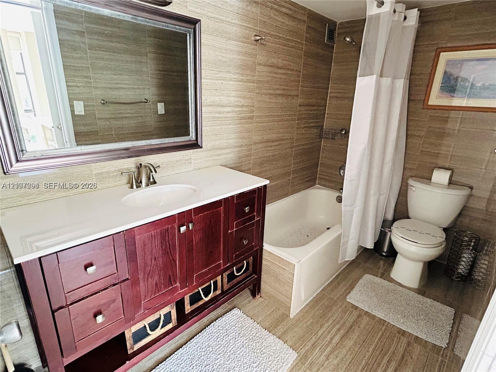 Bathroom featuring a vanity with wood-finish cabinetry, white countertop with integrated sink, framed wall mirror, and floor-to-ceiling tile surrounding the tub and shower combination