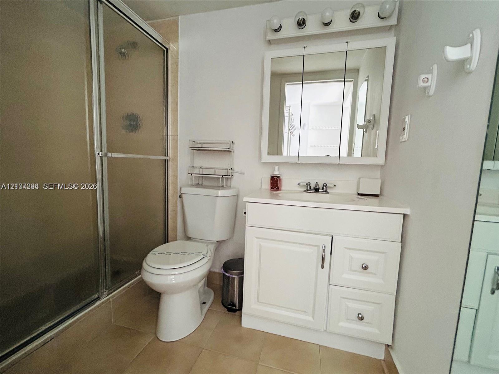 Bathroom featuring a glass-enclosed shower, white vanity cabinet with stone-surfaced countertop, integrated sink, and triple-door medicine cabinet with integrated lighting