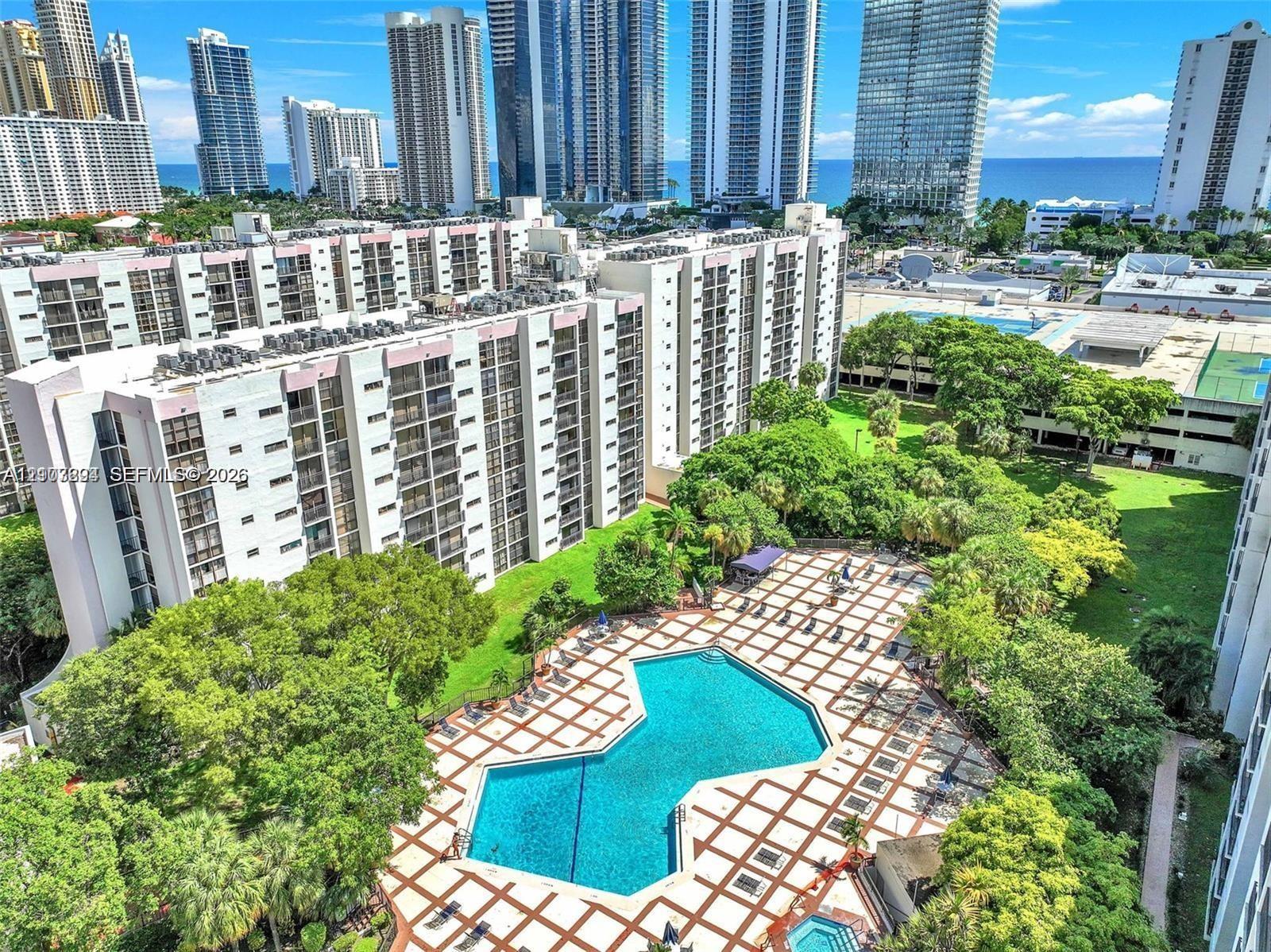Expansive swimming pool with geometric tile surround, lush landscaped grounds, mature trees, and high-rise skyline backdrop