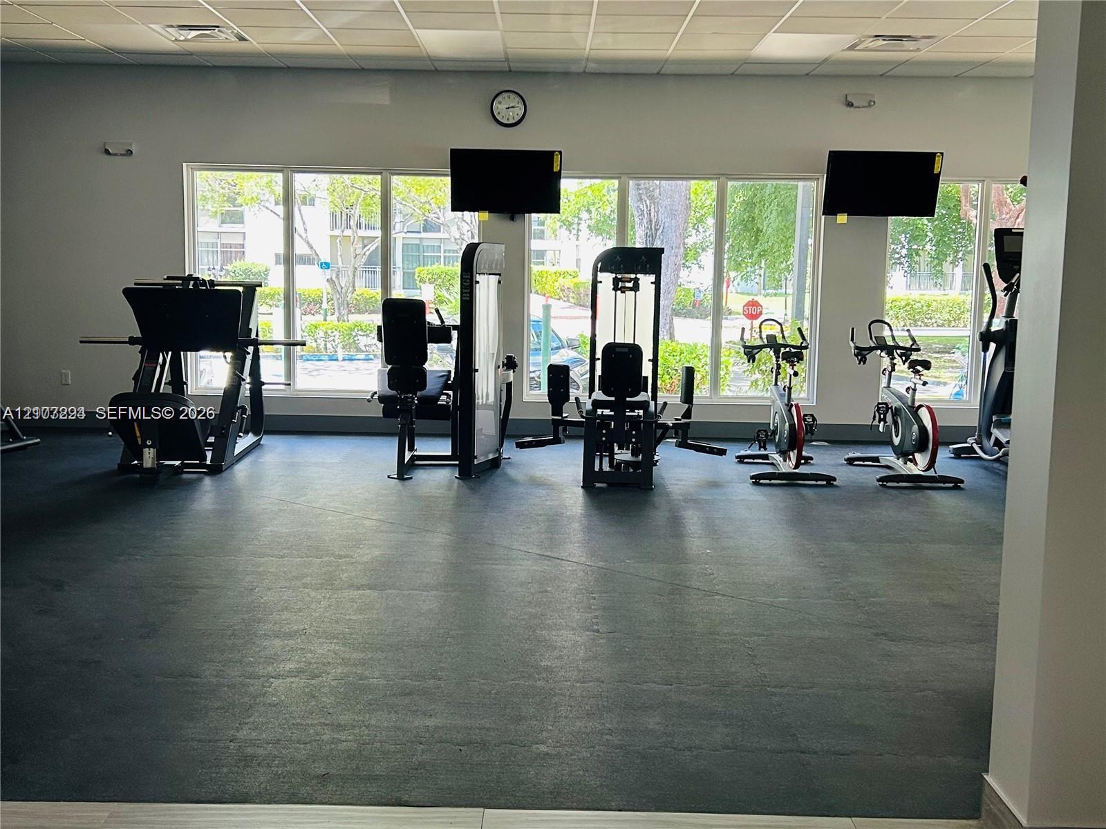 Fitness center featuring durable rubber flooring, a drop-tile ceiling, and oversized windows providing natural lighting